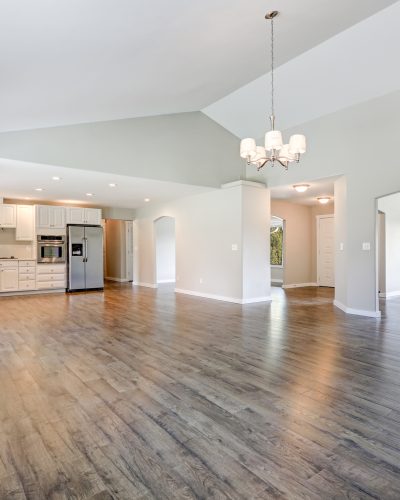 Spacious rambler home interior with vaulted ceiling over glossy laminate floor. Empty light filled dining or living space adjacent to new white kitchen room features pale grey walls. Northwest, USA